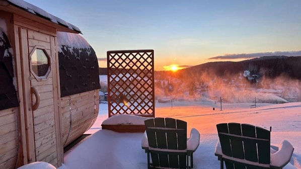 A wooden cabin with a snow-covered roof, a lattice screen, and two Adirondack chairs facing a snowy landscape with a sunset.