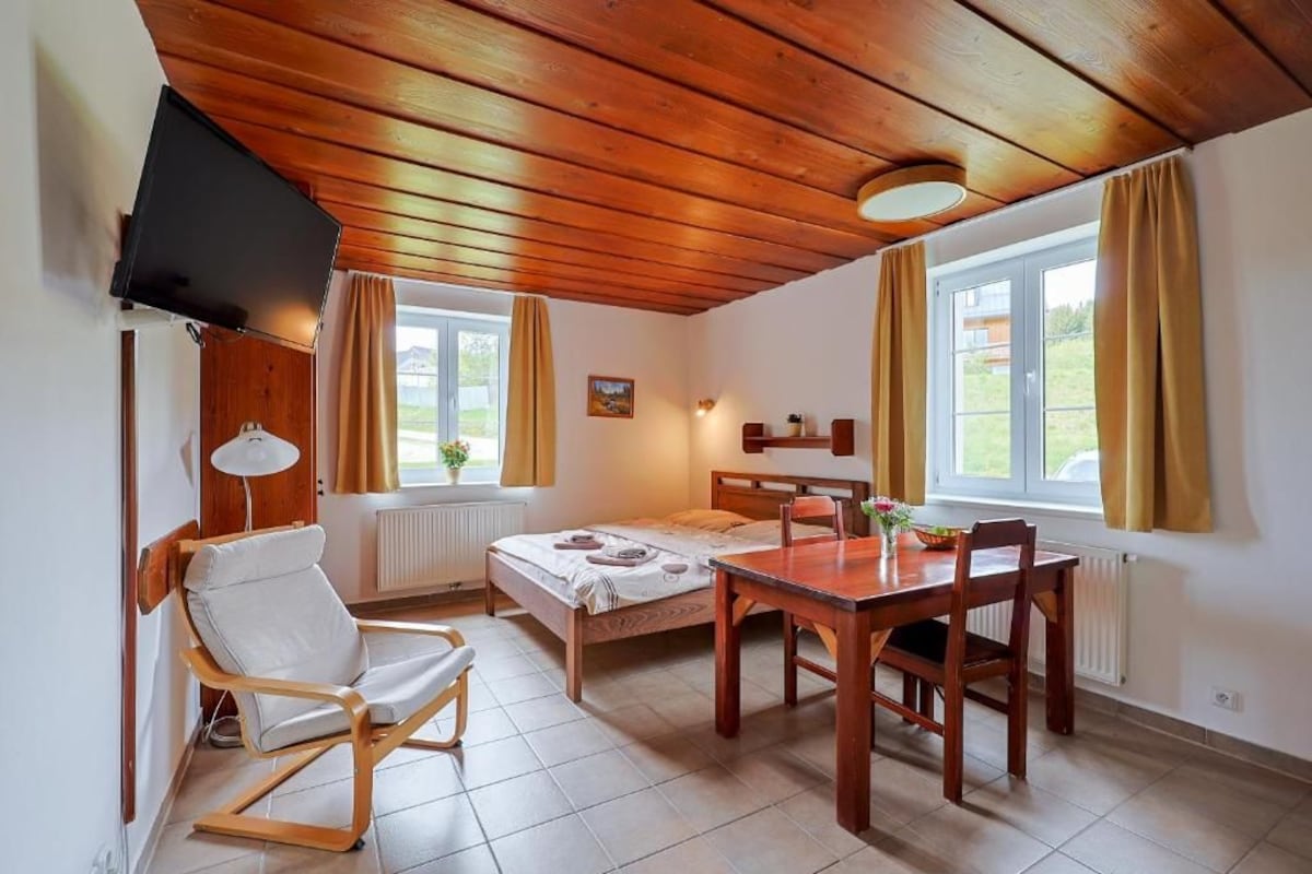 Una stanza con un letto, un tavolo da pranzo in legno con sedie, una poltrona bianca, una televisione a parete e una finestra con tende.