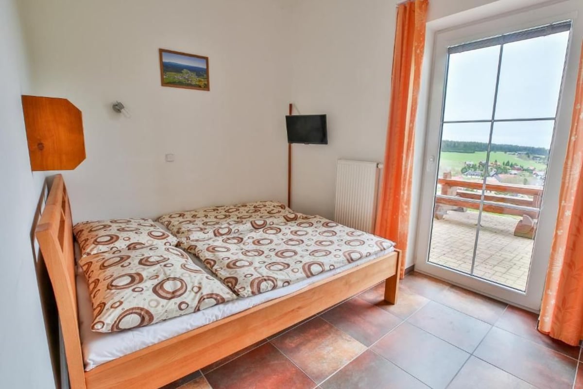 Un letto singolo con copriletto decorato, una televisione a parete, una finestra con tende e vista sull'esterno.