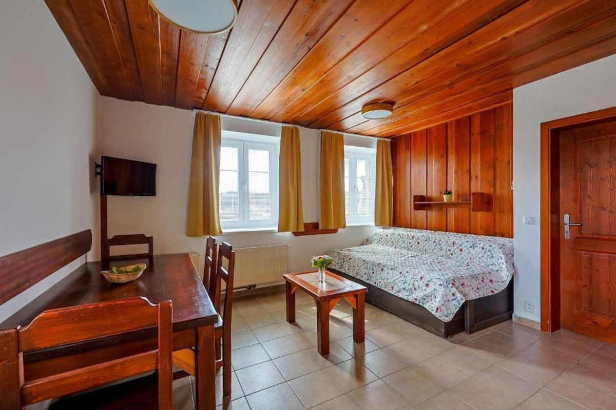 Una stanza con soffitto in legno, un letto con copriletto floreale, un tavolo da pranzo con sedie, una televisione a parete e una finestra con tende.