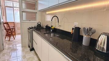 Una cocina con encimera negra, armarios blancos, un lavamanos de acero inoxidable y una ventana con persianas.
