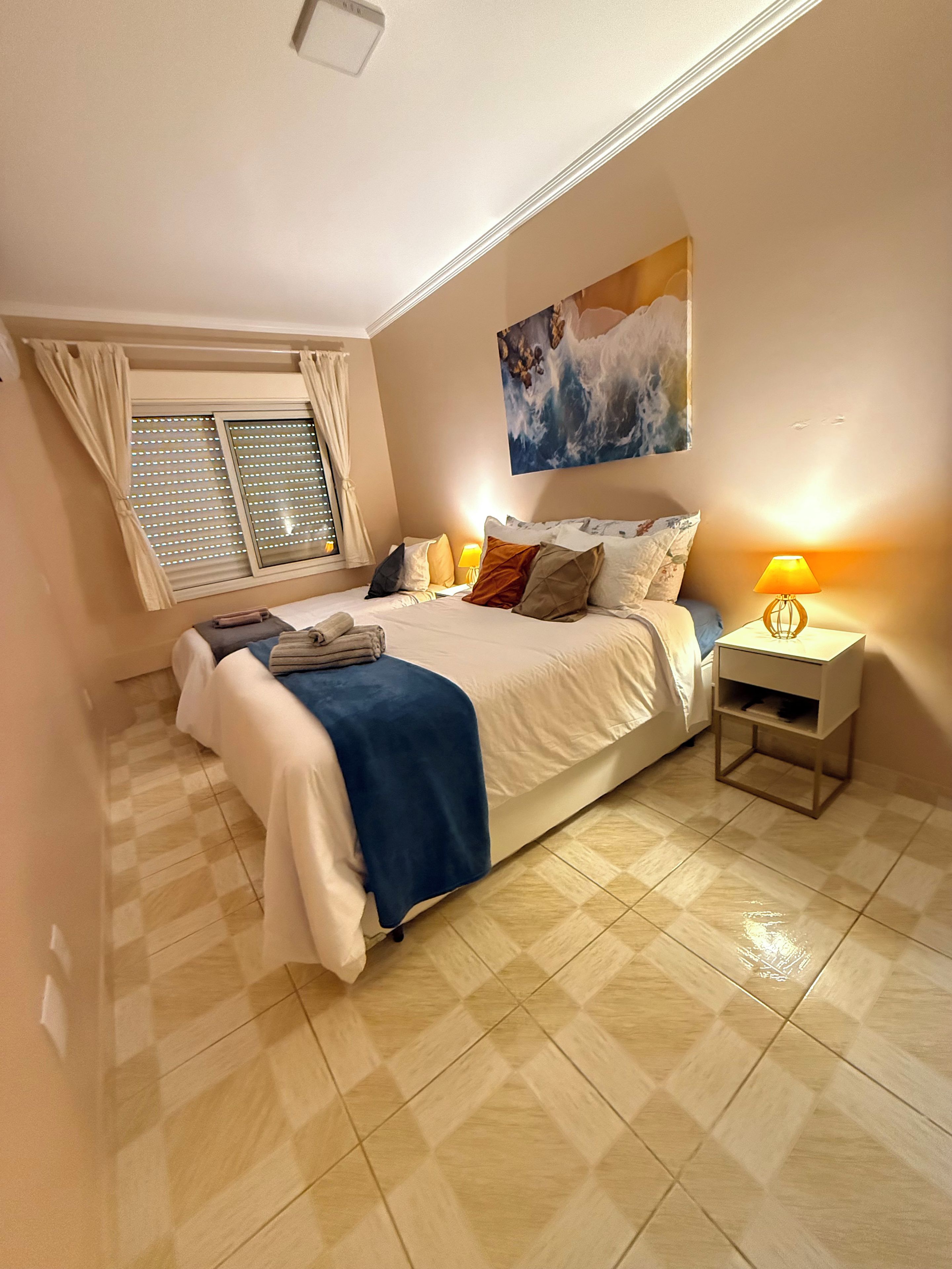 Un dormitorio con cama, mesita de noche, ventana con cortinas, un cuadro en la pared y piso de baldosas.