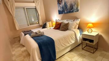 Un dormitorio con cama, mesita de noche, ventana con cortinas, un cuadro en la pared y piso de baldosas.