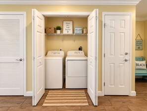 A laundry room with two washing machines, a shelf with laundry supplies, and a door leading to another room. - Luxury Coastal Condo, Pool, Hot Tub, 11th Floor (Panama City Beach)