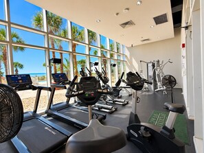 Une salle de sport équipée de tapis de course, de vélos d’appartement et d’appareils de musculation, de grandes fenêtres offrant une vue sur des palmiers et un ciel dégagé.