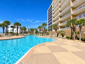 Une piscine dotée d’une terrasse carrelée, entourée de palmiers et bordée d’un immeuble à plusieurs étages avec des balcons.