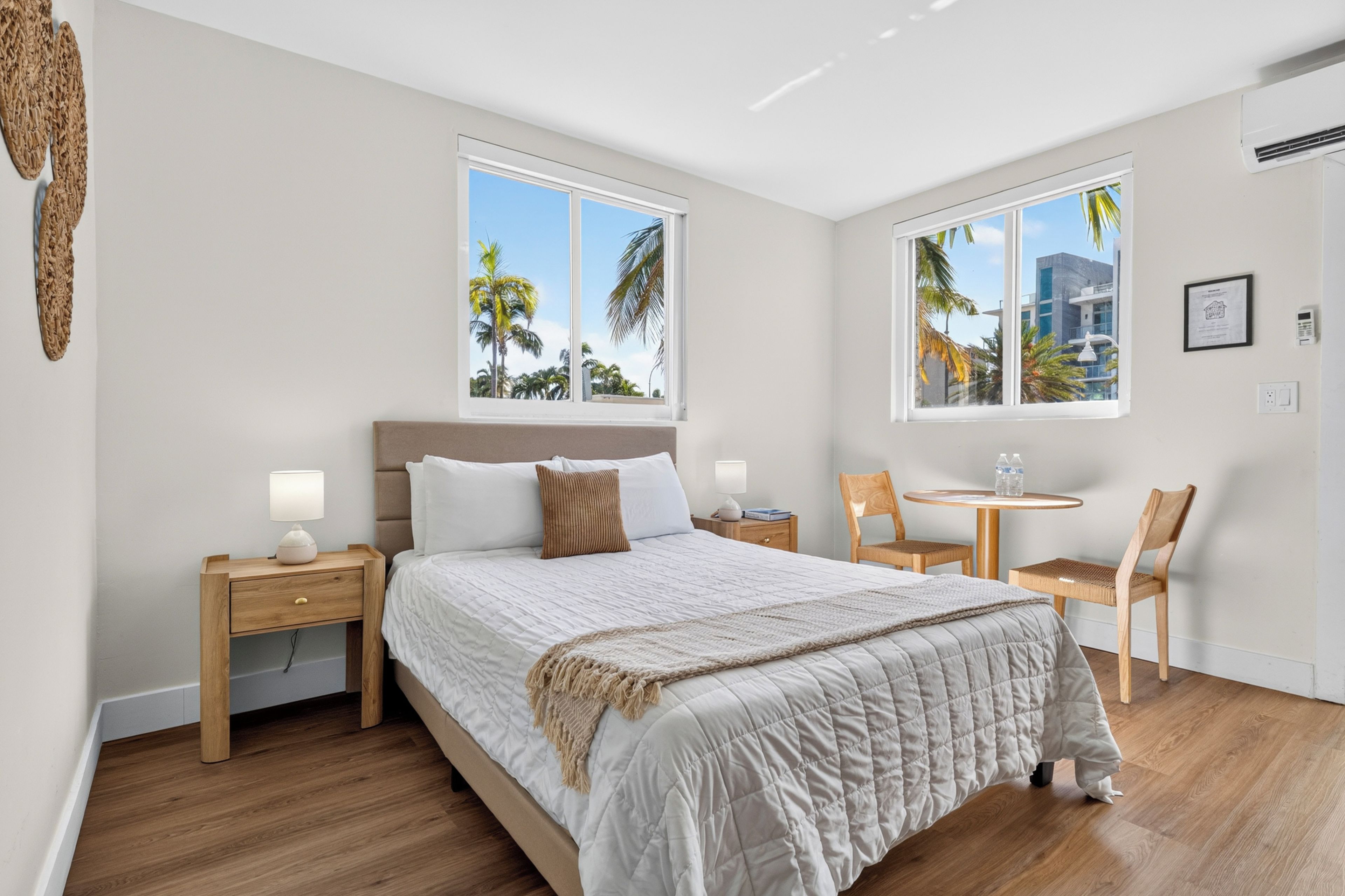 A hotel room with a bed, two bedside tables, a chair, a small table, and two windows with a view of palm trees.