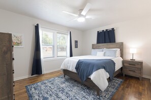 Une chambre à coucher comprenant un lit, une table de chevet, un ventilateur de plafond, une fenêtre avec des rideaux et une porte.