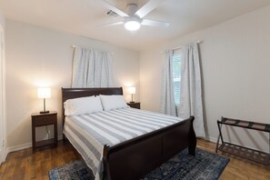 Un dormitorio con una cama, dos mesitas de noche, un ventilador de techo y una ventana con cortinas.