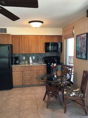 A kitchen with wooden cabinets, a microwave, a refrigerator, and a dining area with a glass table and wicker chairs. - New Lahaina Shores 5th Floor  2 Lanai's Ocean view & Mountain View Corner unit (Lahaina)