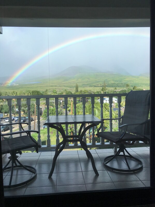 Ein Balkon mit Tisch und Stühlen, mit Blick auf eine Landschaft mit Regenbogen und sanften Hügeln.
