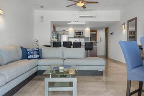 A modern living room with a sofa, a coffee table, and a blue chair. There is a kitchen in the background with a microwave and a refrigerator. - Vista La Jolla 406 | 2BR Suite Ocean View (San José del Cabo)