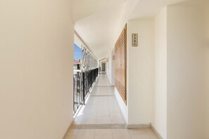 Un couloir avec une porte en bois sur la droite, une rambarde en métal sur la gauche, et un ciel dégagé visible à travers la fenêtre.
