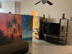 Ein Wohnzimmer mit Flachbildfernseher, einer Couch, einem Deckenventilator und einer Wand mit Palmenmotiv.