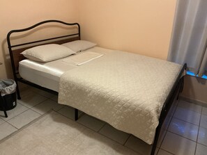 Ein Einzelbett mit Metallgestell, einer weißen Matratze und einer gesteppten Decke. Eine Mülltonne befindet sich links neben dem Bett.