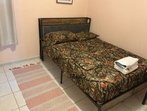 Un lit simple avec une couvre-lit et des oreillers à motifs de camouflage, une tête de lit en bois et une serviette blanche pliée posée sur le pied de lit.