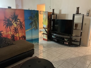 Ein Wohnzimmer mit einer Couch, einem Fernseher auf einem Schrank und Wandbildern mit tropischem Motiv.