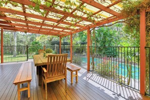 A wooden deck with a table and chairs, a pool, and a pergola with vines. - Moreton Bay Acreage Retreat Pool & Fireplace (Ransome)