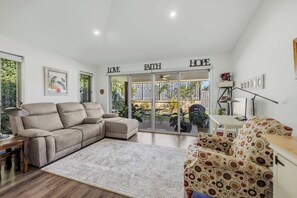 Ein Wohnzimmer mit einem beigen Sofa, einem gemusterten Sessel, einem Couchtisch und einem Esstisch. Ein großes Fenster bietet einen Blick ins Freie.