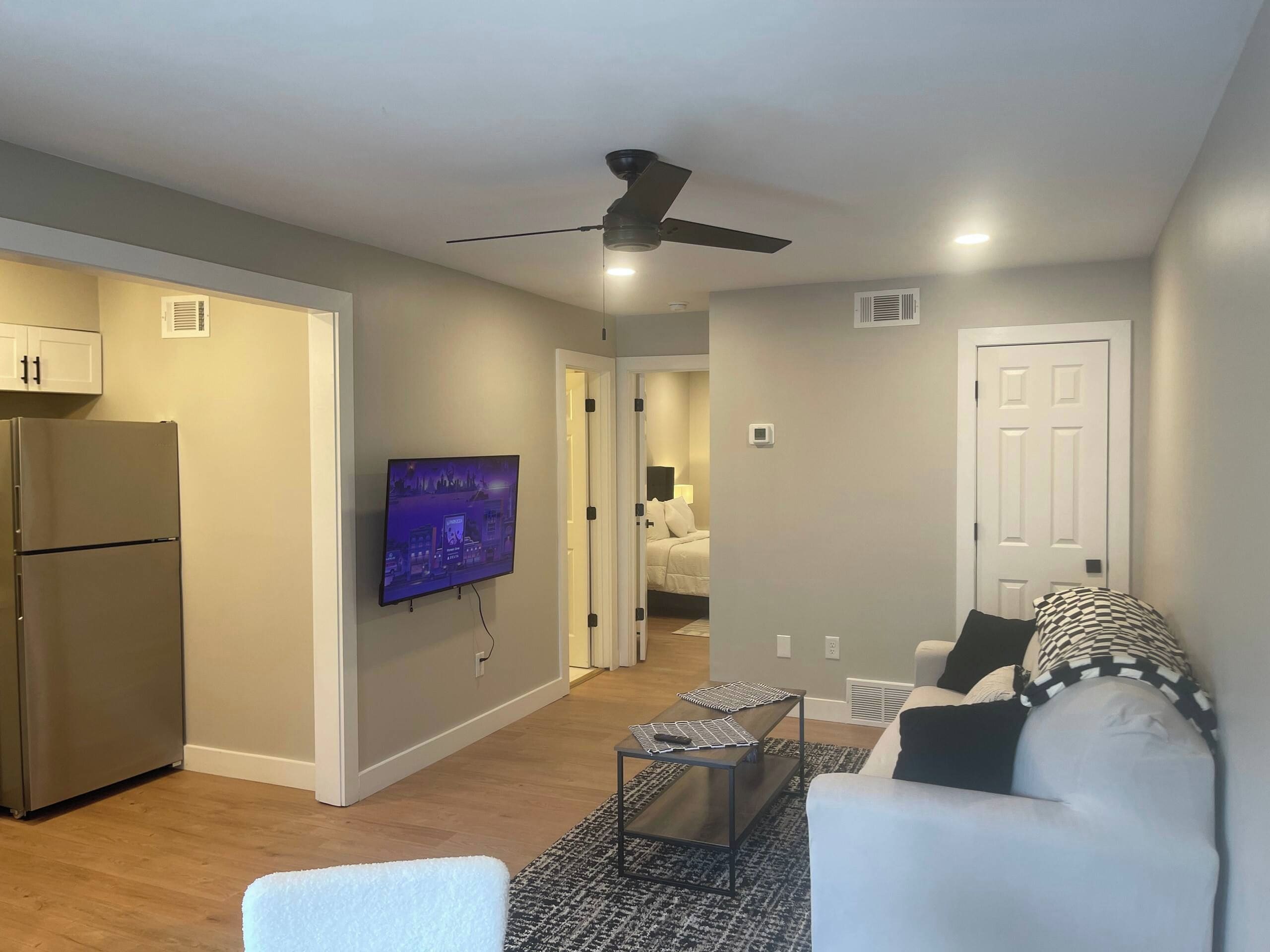 A modern living room with a flat-screen TV, a ceiling fan, and a refrigerator.