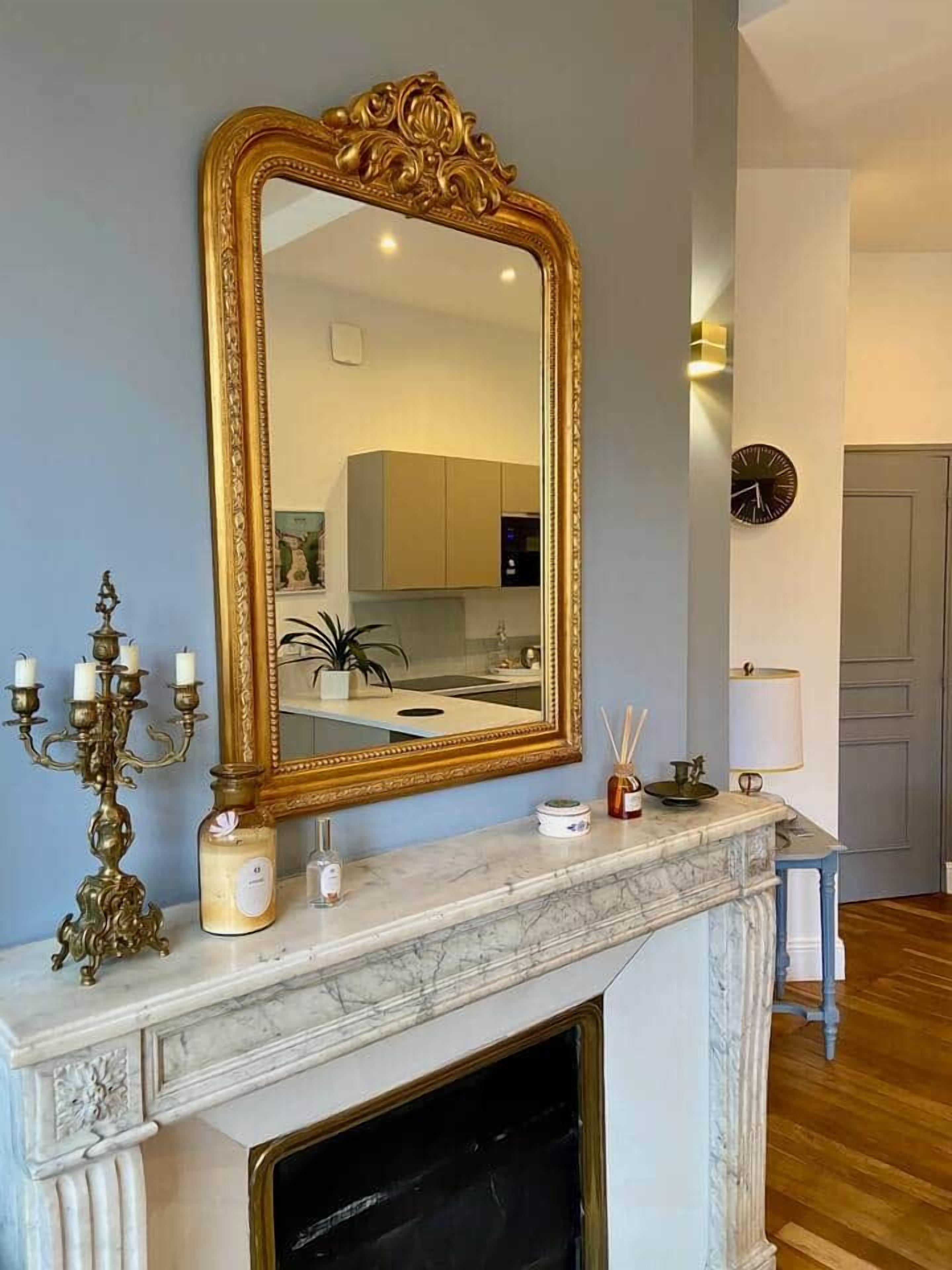 A room with a large ornate mirror, a marble fireplace, a candelabra, and a clock on the wall.