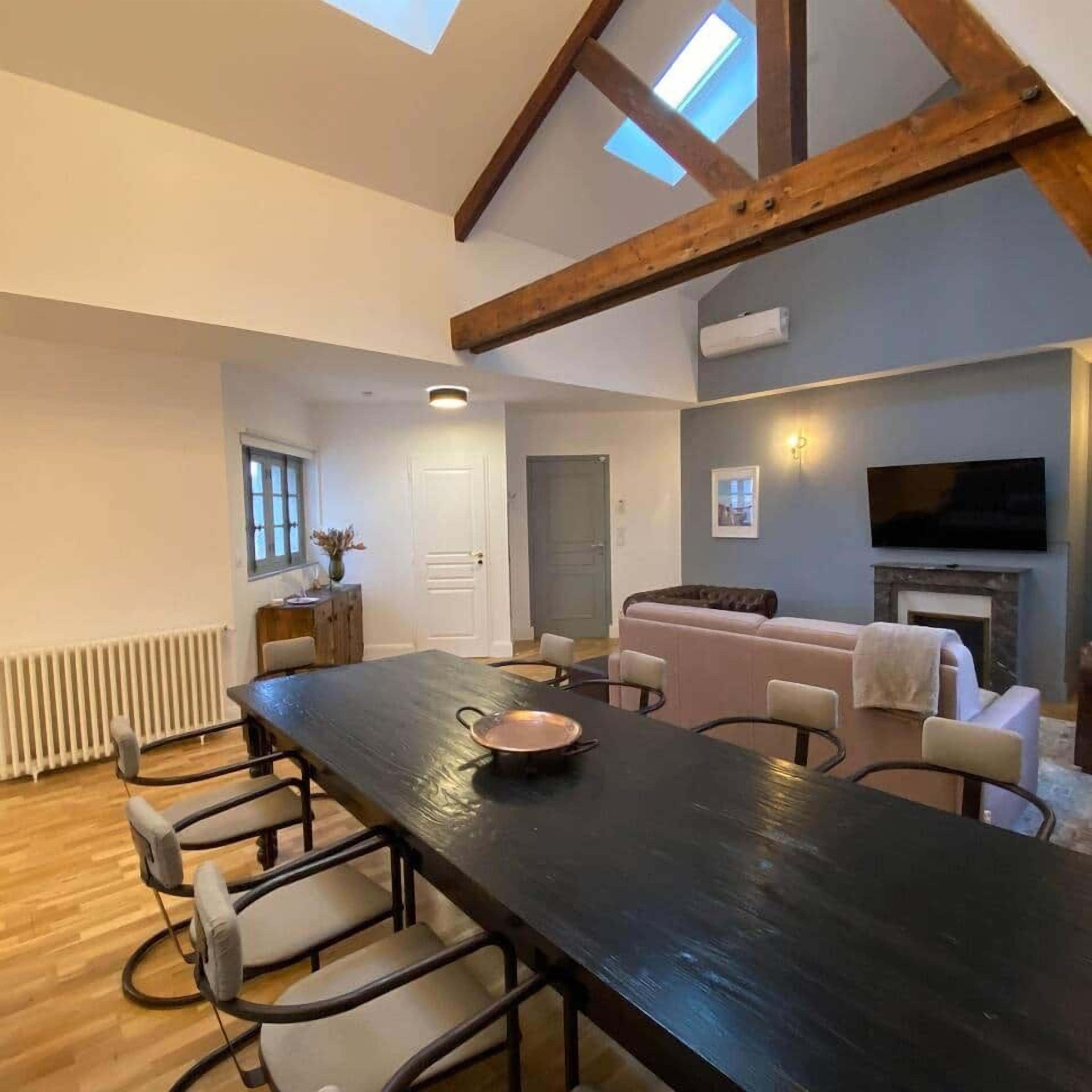 A spacious living area with a long dining table, comfortable seating, a fireplace, and a television mounted on the wall.