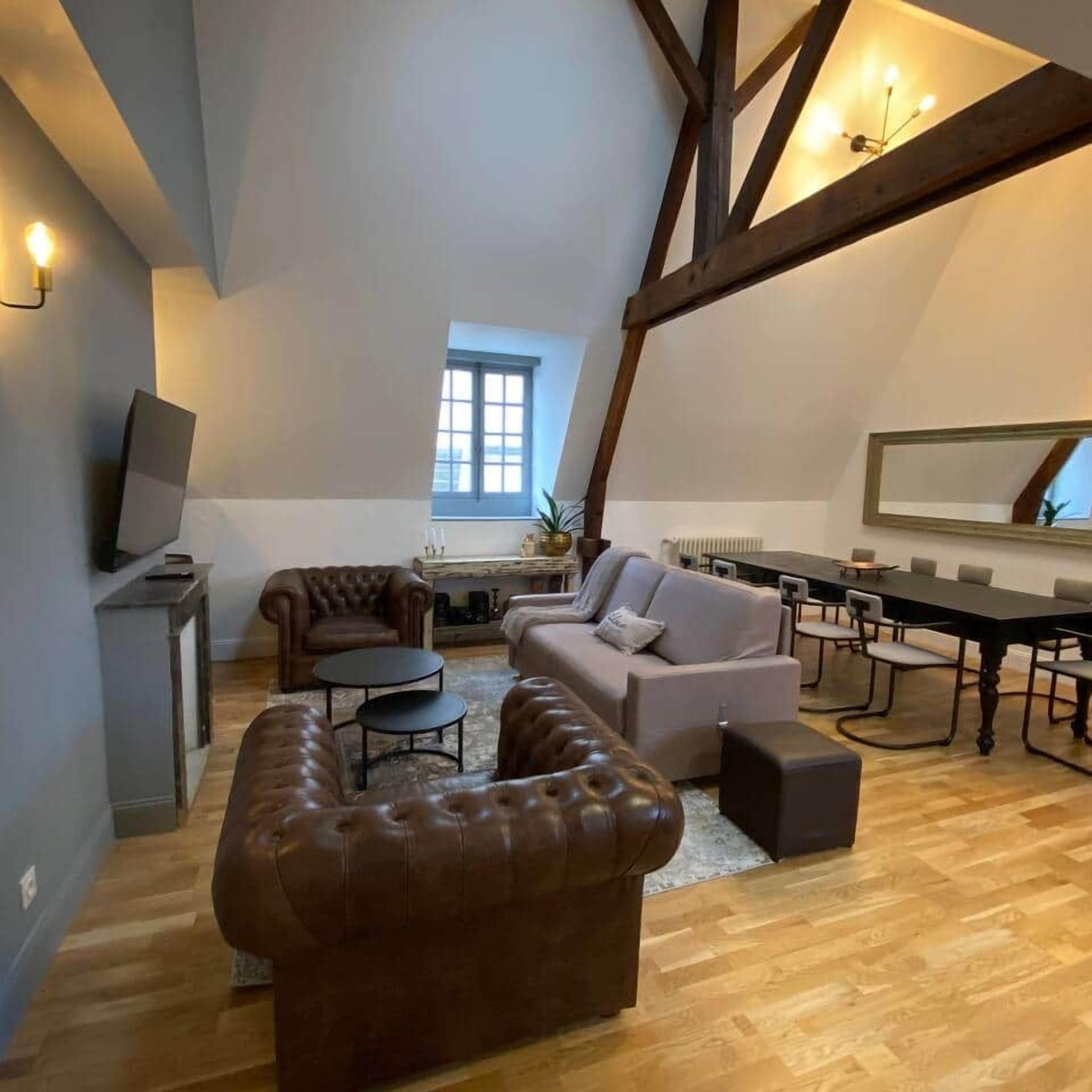 A loft-style living room with a high ceiling, exposed wooden beams, a fireplace, a television, and a dining area.