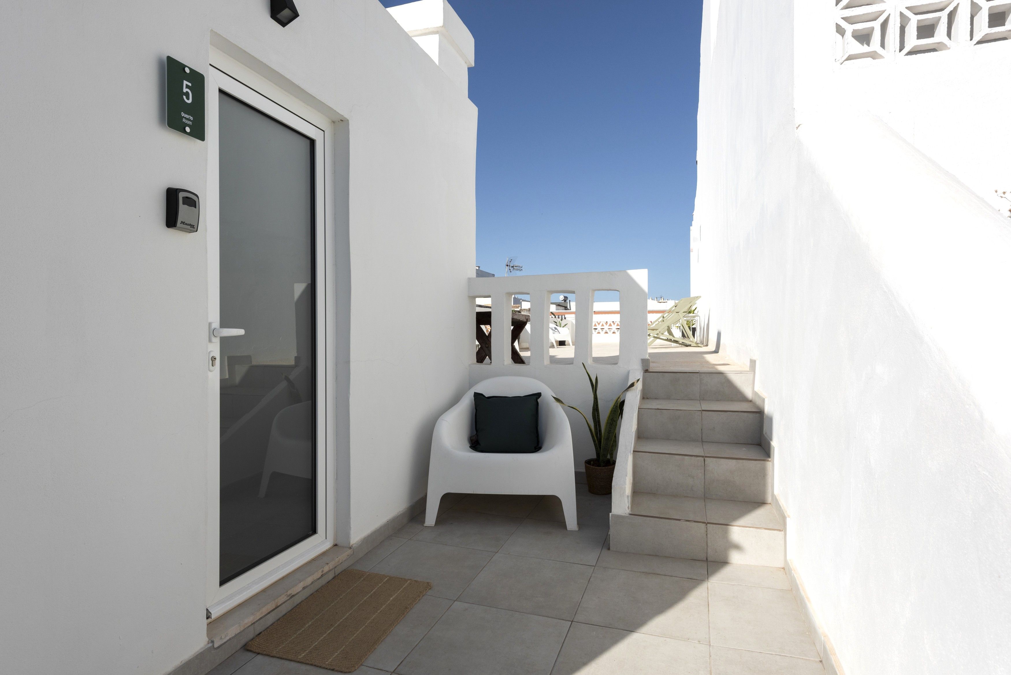 Un bâtiment à l’extérieur blanc, avec une porte portant le numéro « 5 », une petite chaise blanche, une plante en pot et un escalier menant à un étage supérieur.