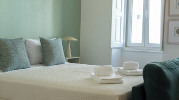 A neatly made bed with pillows, a lamp, and towels on a white bedspread in a room with a window and a green wall.