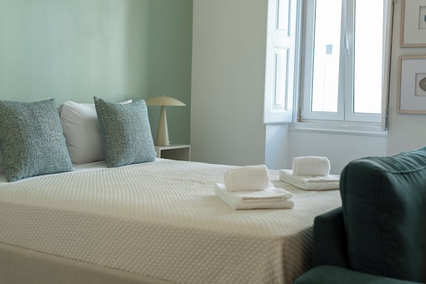 A neatly made bed with pillows, a lamp, and towels on a white bedspread in a room with a window and a green wall.