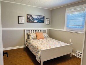 A bedroom with a bed, a window with blinds, and a wall-mounted picture. - Parkwood Nest | 2BR Home Base for Cape Breton (Sydney)