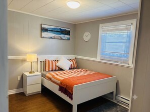 A bedroom with a bed, nightstand, lamp, wall clock, window with blinds, and a painting on the wall. - Parkwood Nest | 2BR Home Base for Cape Breton (Sydney)