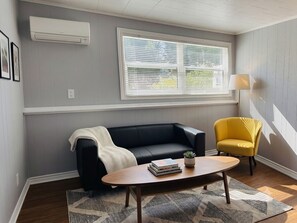 A living room with a black sofa, a yellow armchair, a coffee table with books, a window with blinds, and an air conditioning unit. - Parkwood Nest | 2BR Home Base for Cape Breton (Sydney)