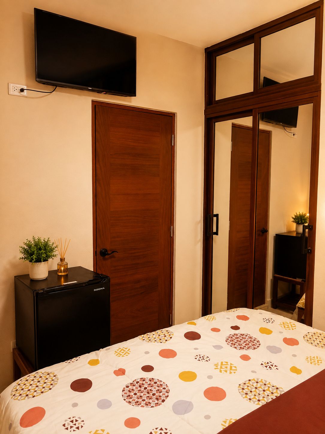 Un dormitorio con cama, televisor montado en la pared, armario de madera y un pequeño mueble negro.