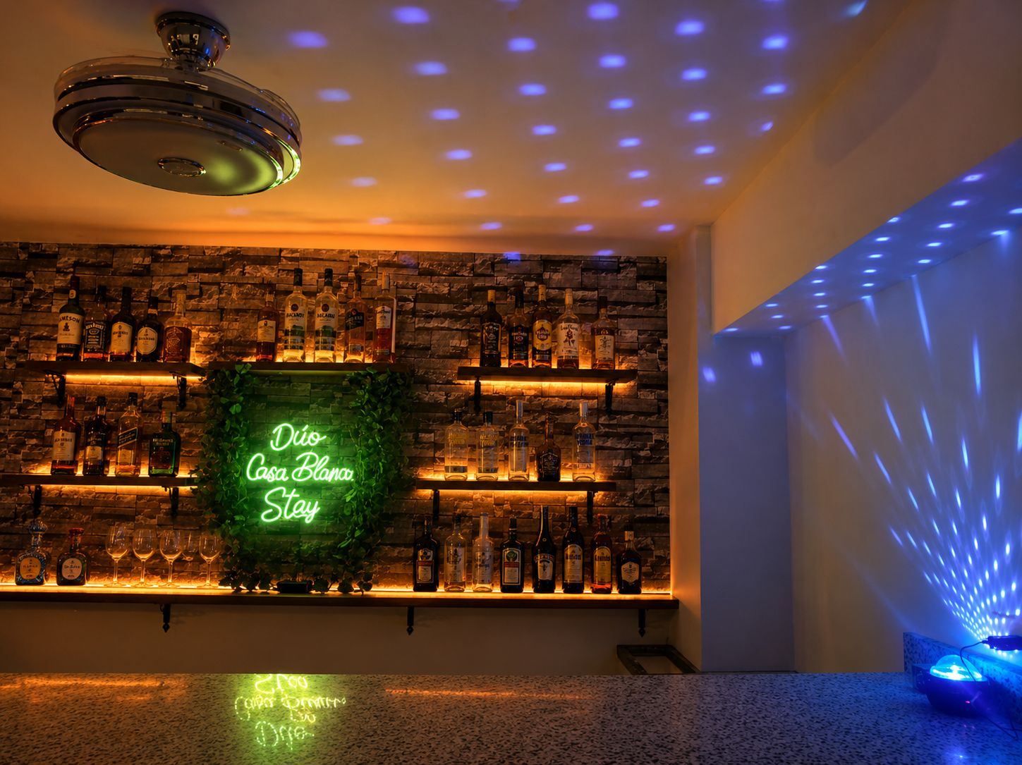 Zona de bar con un letrero de neón, estantes llenos de botellas de licores y una lámpara en el techo.