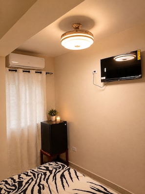 A bedroom with a bed, a ceiling fan, an air conditioning unit, a television mounted on the wall, and a small table with a plant and candle.