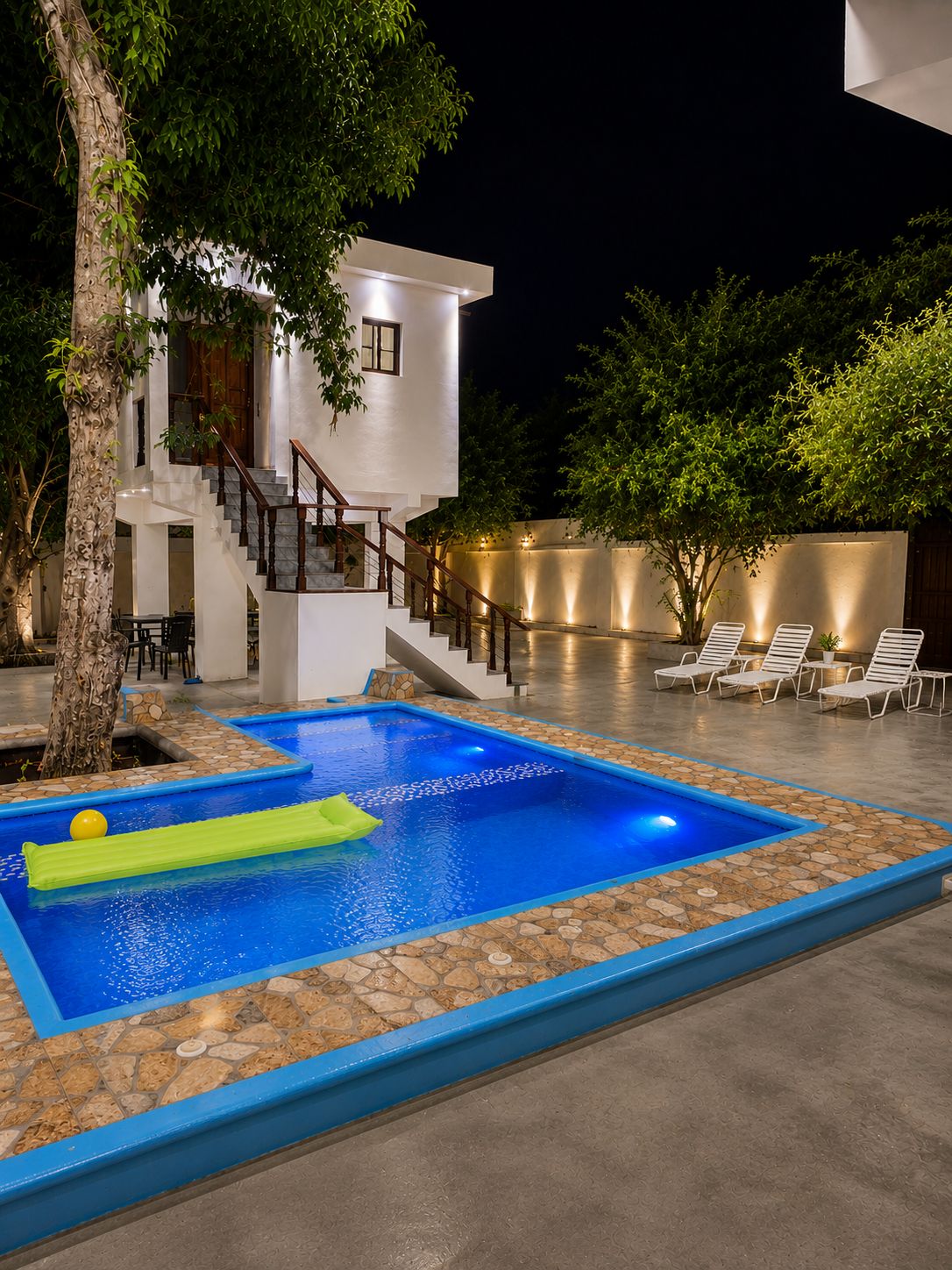 Una piscina moderna al aire libre con borde de piedra, una escalera que lleva a un edificio blanco y sillones de descanso bajo árboles.