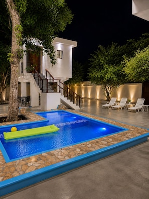 A modern outdoor swimming pool with a stone border, a staircase leading to a white building, and lounge chairs under trees.