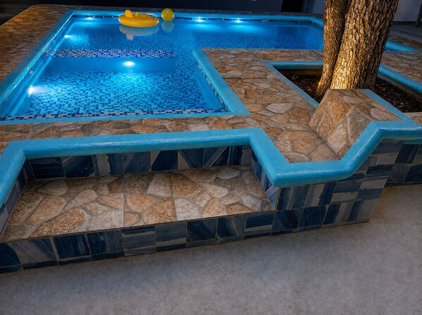 A swimming pool with a tiled deck and a tree nearby.