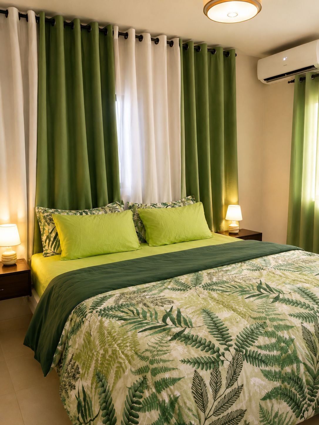 Ein Bett mit einer Bettdecke in Grün-Weiß mit Blätermotiv, zwei grüne Kissen und ein Nachttisch mit einer Lampe.