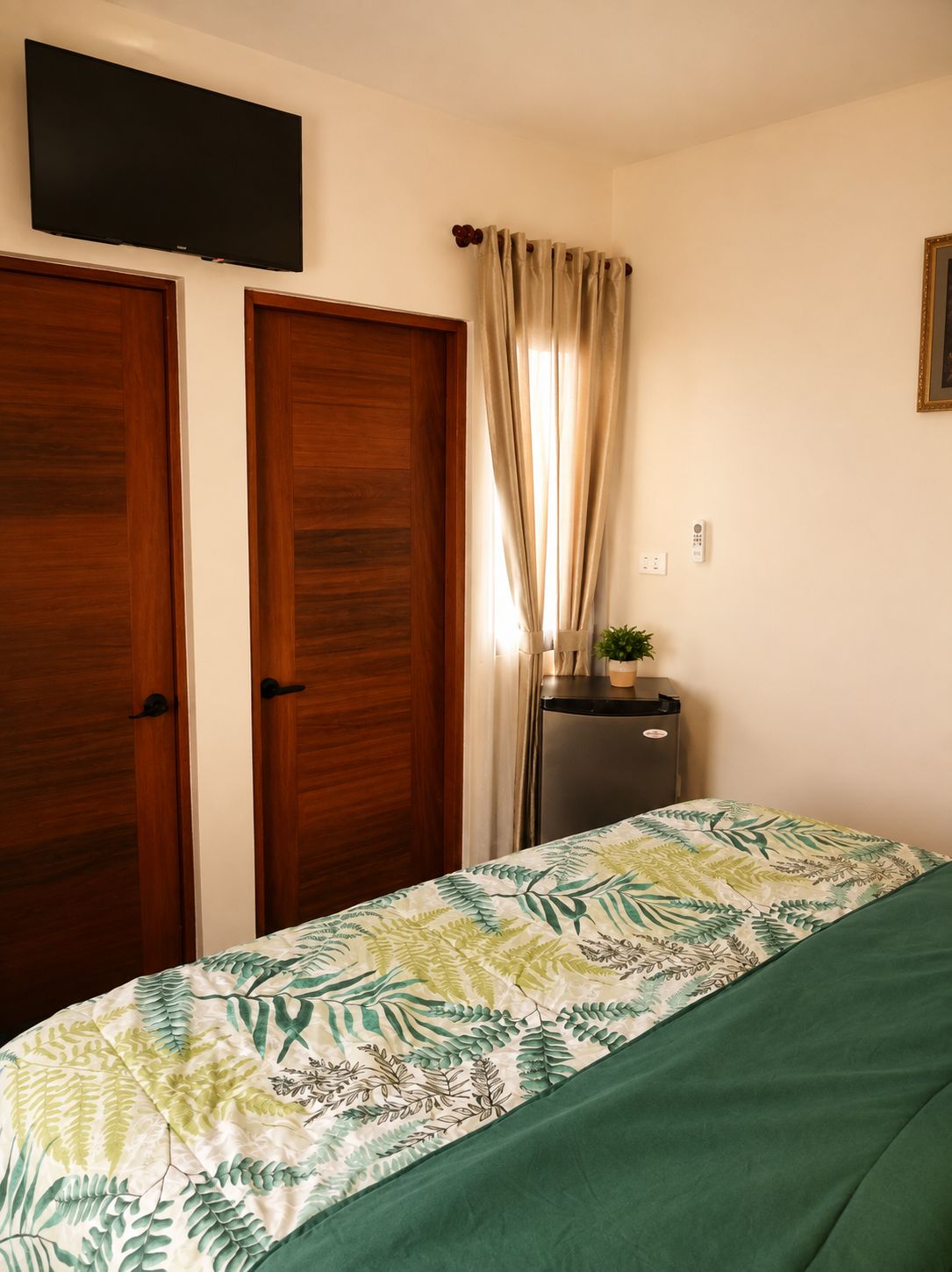 Un dormitorio con dos puertas de madera, una cama con colcha floral, una mesita con una planta y un televisor montado en la pared.