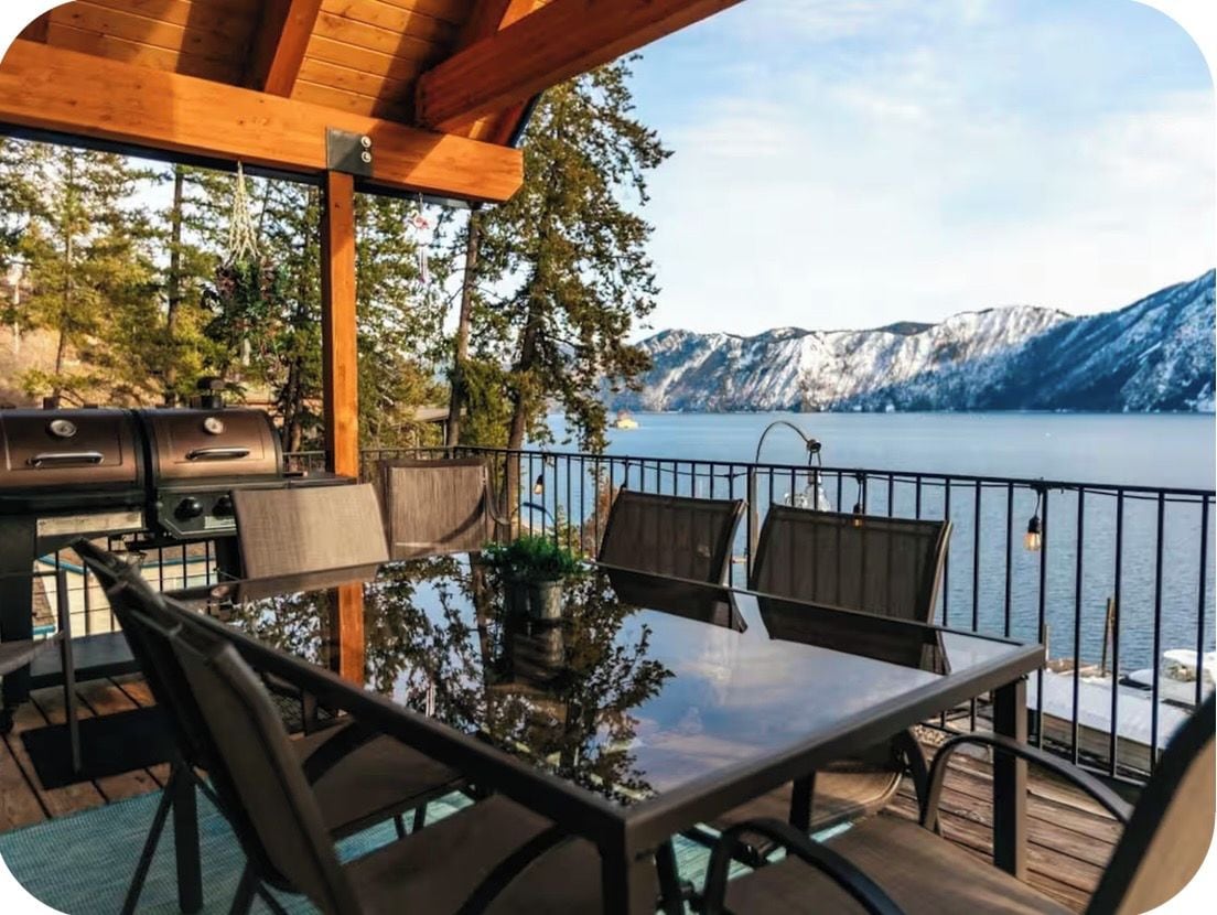 An outdoor dining area with a view of a lake and mountains, featuring a grill and seating.