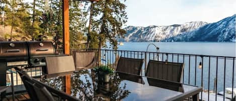 An outdoor dining area with a view of a lake and mountains, featuring a grill and seating.