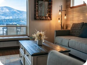 A cozy living room with a view of a mountain, a wooden coffee table with a vase of dried flowers, a sofa, and a wall-mounted light fixture. -  Waterfront Home in Bayview with private boat dock on Lake Pend Oreille (Bayview)