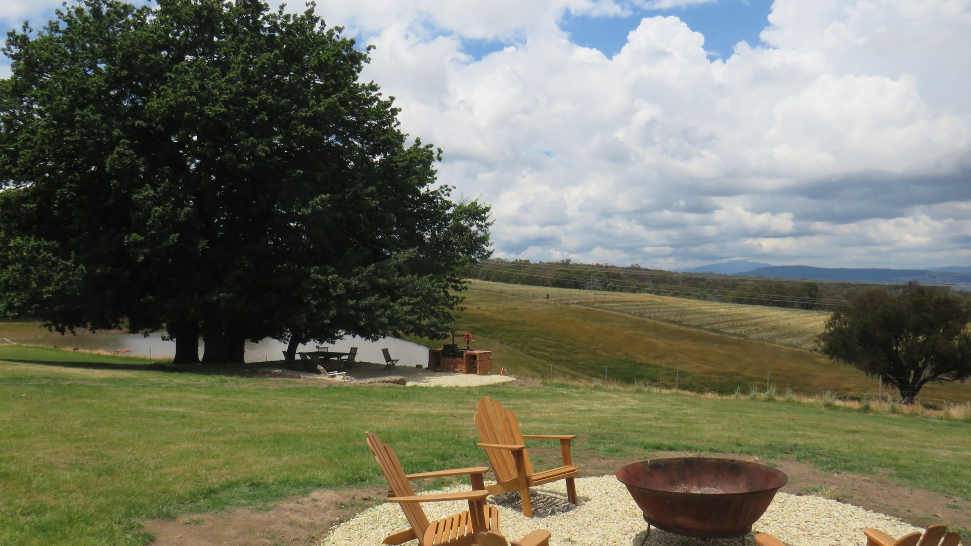 Een terras aan het water met houten stoelen en een vuurkorf, omringd door een grasveld en een grote boom.