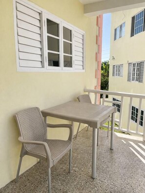 A balcony with a table and chairs, a window with shutters, and a building with balconies in the background. - Modern Refurbed Condo | Resort Pool | Sleeps 4 (Ocho Rios)