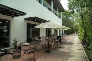 Ein Straßencafé mit Tischen, Stühlen und Sonnenschirmen, sowie ein Gebäudeeingang.