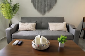 Ein graues Sofa mit zwei Kissen, einer Fernbedienung, einer Kerze und einem kleinen Topf mit Pflanze auf einem Holztisch.