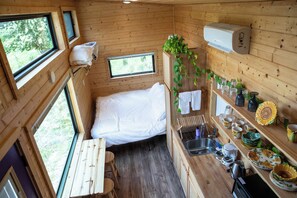 Ein kleines, hölzernes Blockhäuschen mit einem Einzelbett, einer kleinen Küchenzeile und einem Fenster mit Blick auf Grünflächen.
