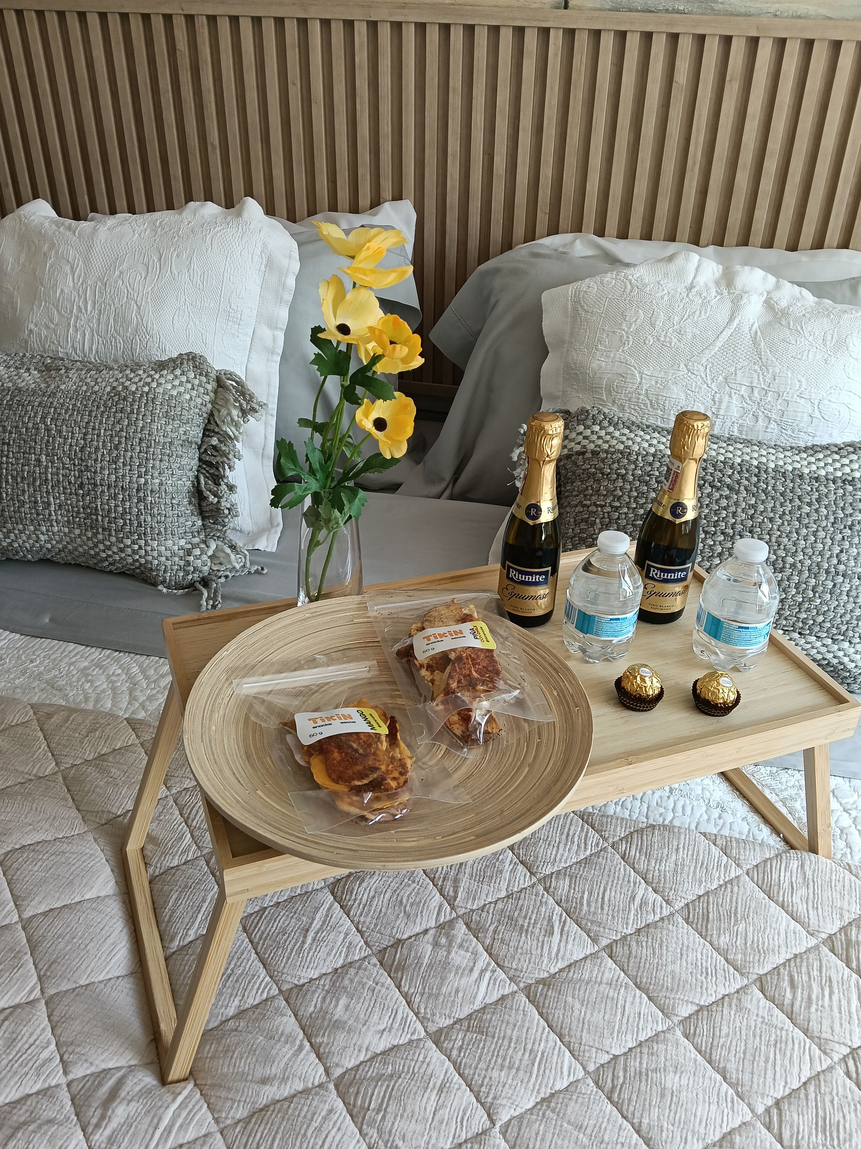 A bed with pillows, a bedside table with a bottle of champagne, water bottles, and packaged snacks.